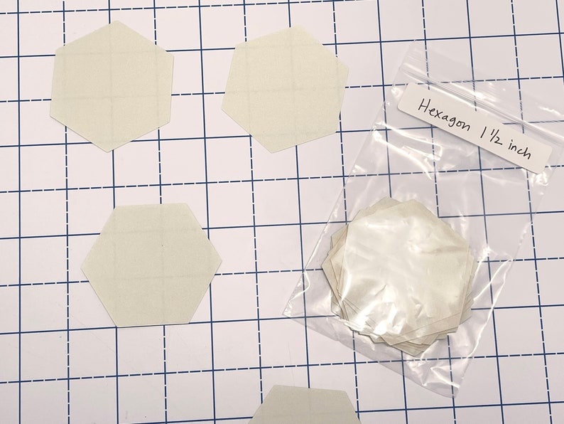 K&ouml;nnte beinhalten: Auf einer karierten Oberfl&auml;che liegen mehrere cremefarbene sechseckige Formen, jeweils etwa 3,8 cm breit. Ein klarer Beutel enth&auml;lt weitere Formen. Ein Etikett tr&auml;gt die Aufschrift "Hexagon 1 1/2 inch".