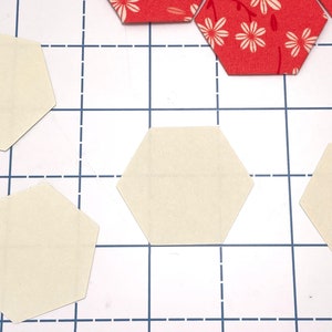 May include: Cream and red floral hexagon fabric pieces are arranged on a white cutting mat with a blue grid. These fabric pieces are for quilting or crafting projects.