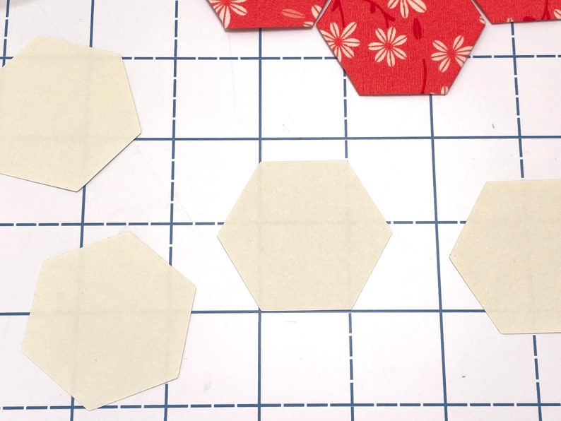 May include: Cream and red floral hexagon fabric pieces are arranged on a white cutting mat with a blue grid. These fabric pieces are for quilting or crafting projects.