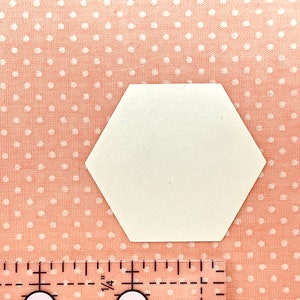 May include: A white, hexagonal paper shape rests on pink fabric dotted with white circles. A clear ruler with black markings and numbers is in the lower portion of the image, measuring in inches.