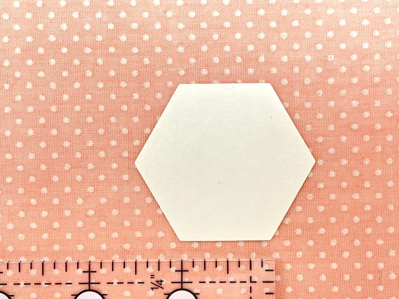 May include: A white, hexagonal paper shape rests on pink fabric dotted with white circles. A clear ruler with black markings and numbers is in the lower portion of the image, measuring in inches.