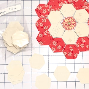 May include: Hexagon-shaped fabric pieces, some in a clear bag labelled "Hexagon 1 inch", are arranged on a grid. Some hexagons are cream-coloured, while others feature a red floral pattern. The pieces are for quilting or crafting, with each hexagon measuring 2.54 cm.