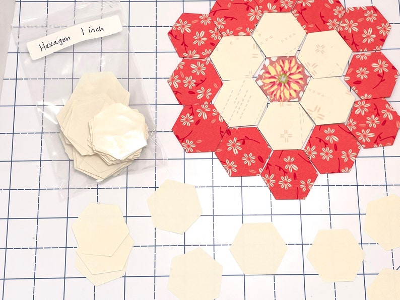 May include: Hexagon-shaped fabric pieces, some in a clear bag labelled "Hexagon 1 inch", are arranged on a grid. Some hexagons are cream-coloured, while others feature a red floral pattern. The pieces are for quilting or crafting, with each hexagon measuring 2.54 cm.