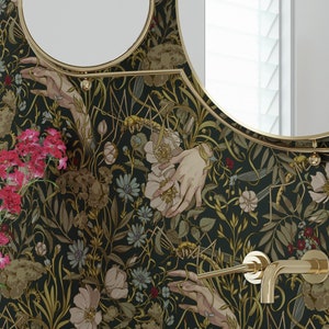 Puede incluir: Escena de ba&ntilde;o con lavabo ovalado blanco, grifos dorados y espejo redondo. El papel pintado presenta un dise&ntilde;o oscuro de flores y manos. Un jarr&oacute;n con flores rosas se encuentra sobre una superficie de madera.
