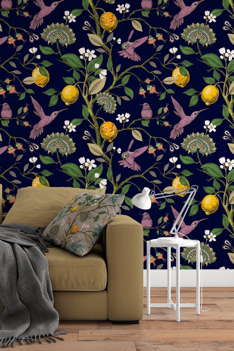May include: A brown couch with a gray throw blanket and a patterned pillow sits in front of a wall with a dark blue background and a repeating pattern of lemons, flowers, and hummingbirds.