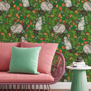 Op de afbeelding: Een roze rieten stoel met een groen kussen staat voor een groene behang met een herhalend patroon van witte konijnen en rode bessen.