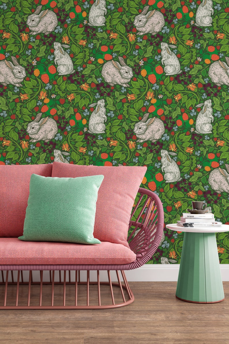 Op de afbeelding: Een roze rieten stoel met een groen kussen staat voor een groene behang met een herhalend patroon van witte konijnen en rode bessen.