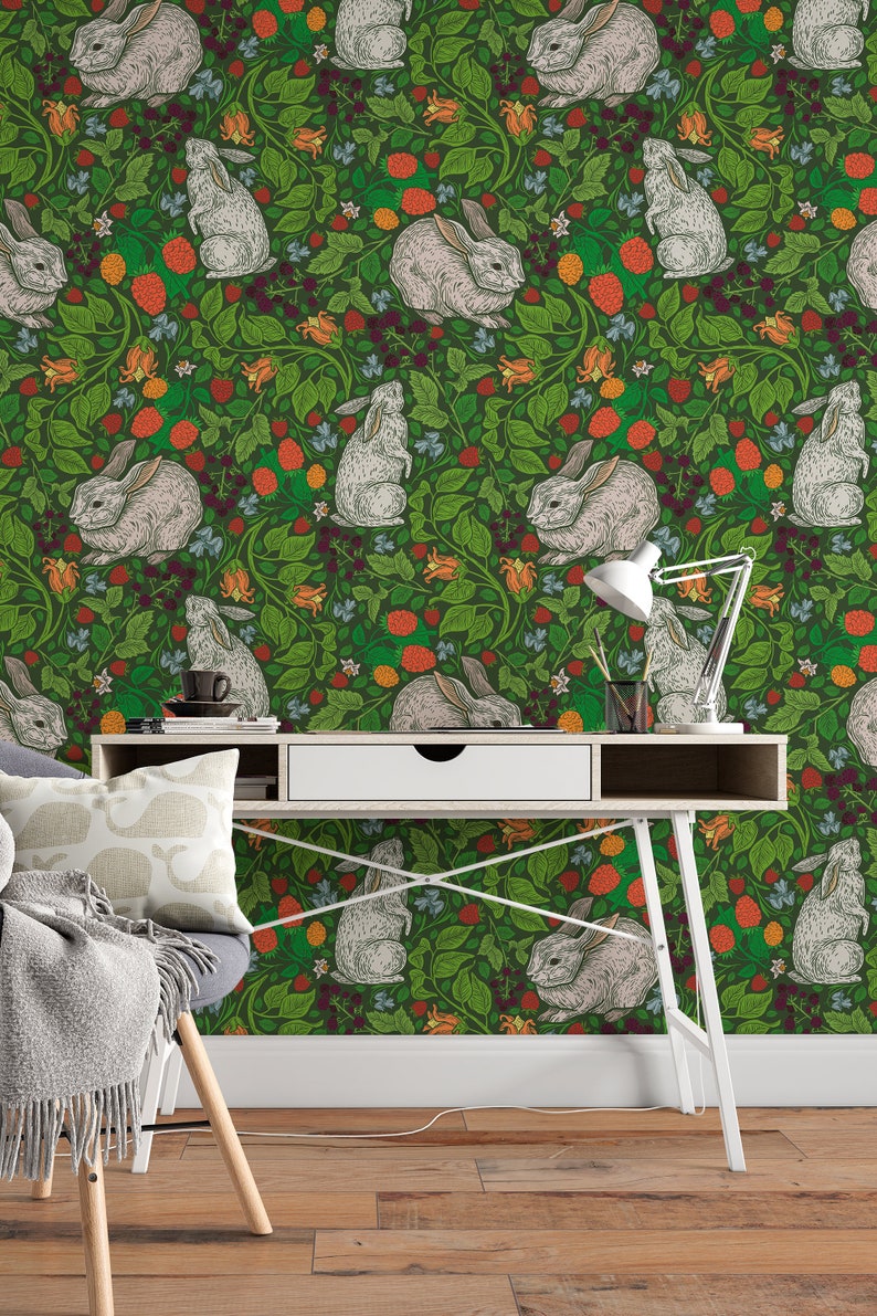 Op de afbeelding: Een groene behang met een herhalend patroon van witte konijnen en rode en oranje bessen. Het behang is achter een wit bureau met een lade en een witte lamp.