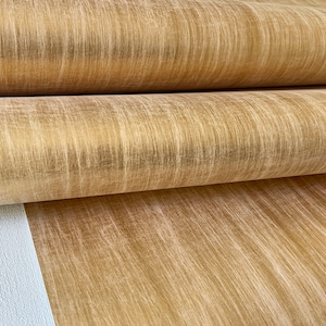 Puede incluir: Un rollo de papel tapiz metálico dorado con una textura cepillada sutil. El papel tapiz tiene un tono dorado cálido y un aspecto ligeramente desgastado.