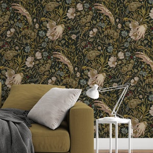 Puede incluir: Papel pintado floral oscuro con manos, flores y follaje en tonos verdes, dorados y crema. Un sof&aacute; color mostaza con una manta gris y una mesita auxiliar blanca con una l&aacute;mpara.