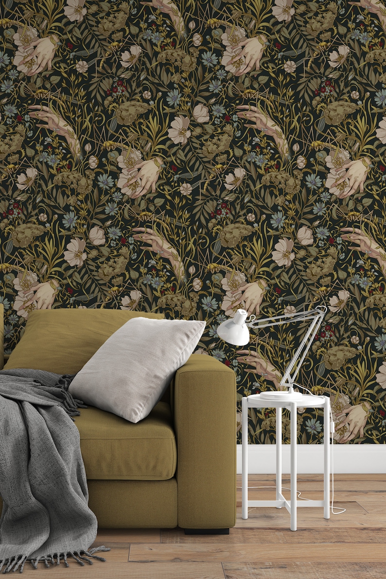 Puede incluir: Papel pintado floral oscuro con manos, flores y follaje en tonos verdes, dorados y crema. Un sof&aacute; color mostaza con una manta gris y una mesita auxiliar blanca con una l&aacute;mpara.