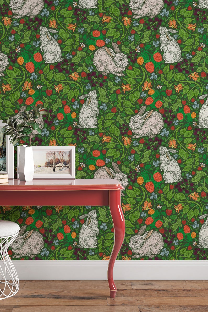 Op de afbeelding: Groene behang met een herhalend patroon van witte konijnen en rode bessen. De behang is te zien in een kamer met een rode tafel en een witte kruk.