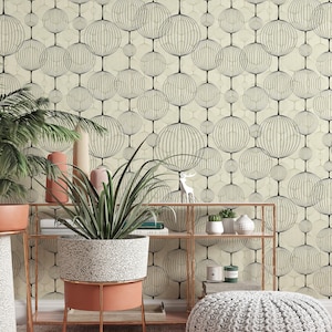 以下が含まれることがあります： ベージュの壁に、黒い円形にグリッド模様が施された幾何学模様の壁紙が貼られた部屋。金色の金属製の棚には、2段の棚があり、鉢植えの植物、本、キャンドルが置かれている。ピンクとベージュの敷物の上に、グレーのニット製のオットマンが置かれている。