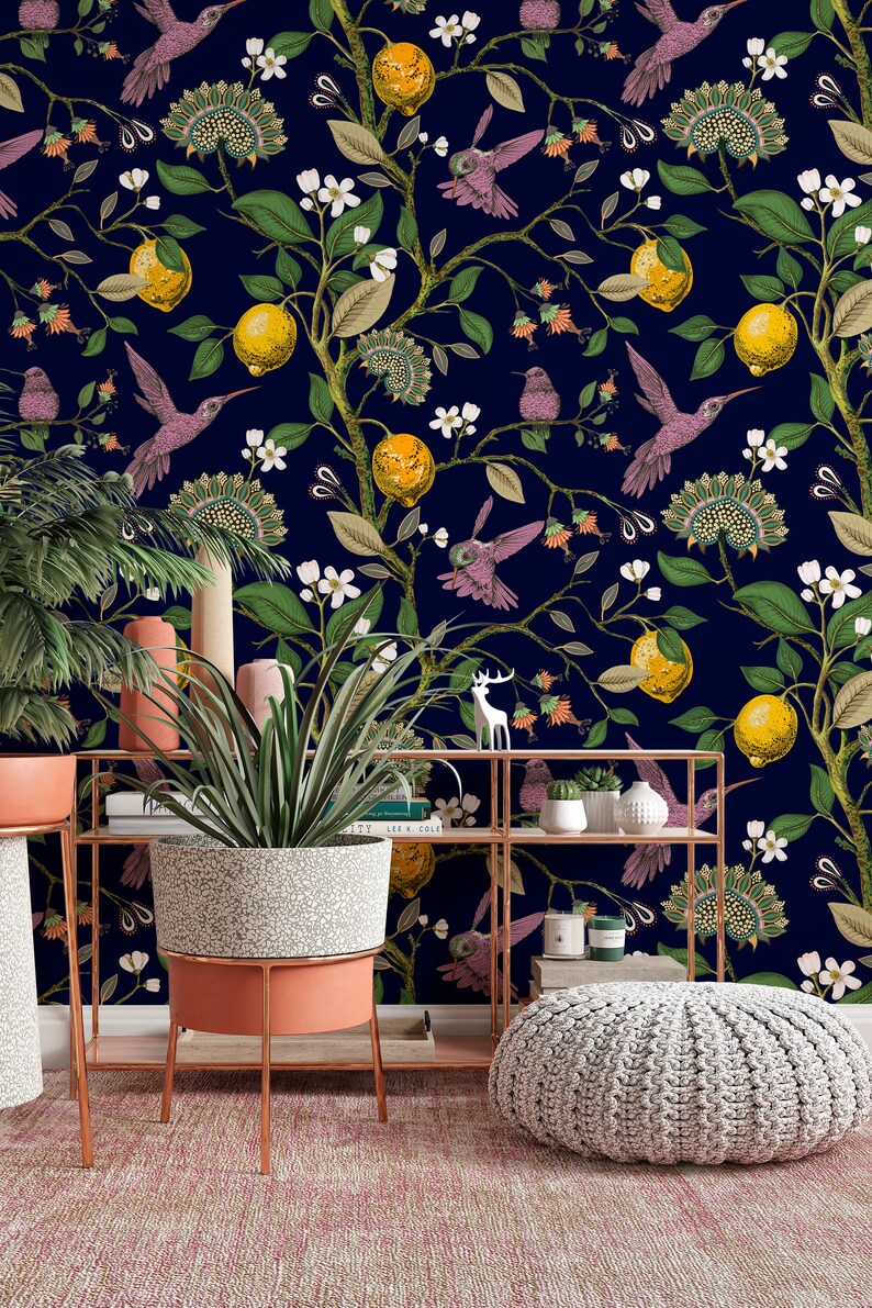 May include: A navy blue wallpaper with a repeating pattern of lemon trees, hummingbirds, and white flowers. The wallpaper is displayed in a room with a potted plant, a shelf, and a decorative footstool.