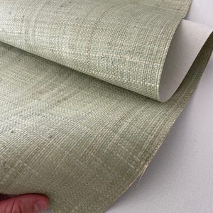 May include: Close-up of a textured, light green woven material, possibly wallpaper or fabric. The material has a natural, organic appearance with visible fibers and a subtle pattern. The back of the material is a smooth, off-white color.