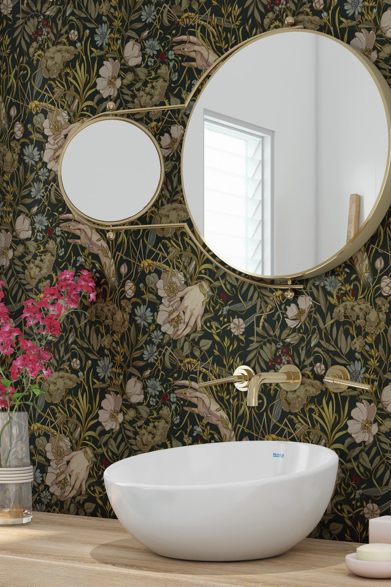 Puede incluir: Escena de ba&ntilde;o con lavabo ovalado blanco, grifos dorados y espejo redondo. El papel pintado presenta un dise&ntilde;o oscuro de flores y manos. Un jarr&oacute;n con flores rosas se encuentra sobre una superficie de madera.