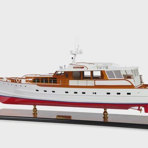 Könnte beinhalten: Ein Modell einer Yacht mit weißem Rumpf, roter Wasserlinie und Holzakzenten. Die Yacht hat eine kleine amerikanische Flagge am Heck und ist auf einem schwarzen Sockel montiert. Das Modell hat ein detailliertes Design mit Fenstern und einem Radarmast.