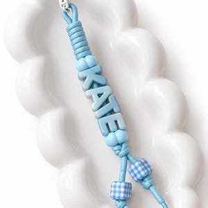 May include: A light blue keyring featuring a white circular clasp and a cord threaded with light blue letter beads spelling KATE. The design includes round light blue beads and two square beads with a white and blue gingham check pattern.