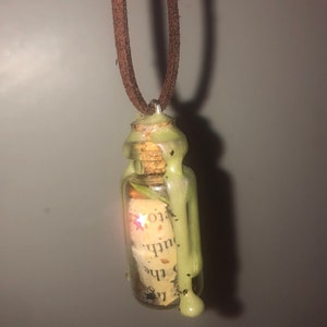 Może przedstawiać: Naszyjnik z zieloną, woskowaną butelką szklaną z korkowym korkiem i zwiniętym kawałkiem papieru w środku. Butelka wisi na brązowym sznurku skórzanym.
