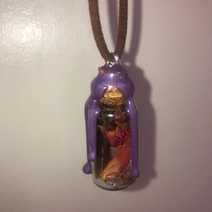Peut inclure: Un pendentif en forme de bouteille en verre scell&eacute; &agrave; la cire violette avec un bouchon en li&egrave;ge et des fleurs s&eacute;ch&eacute;es et des cristaux &agrave; l'int&eacute;rieur, suspendu &agrave; un cordon en daim brun.