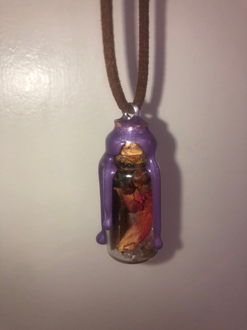 Peut inclure: Un pendentif en forme de bouteille en verre scell&eacute; &agrave; la cire violette avec un bouchon en li&egrave;ge et des fleurs s&eacute;ch&eacute;es et des cristaux &agrave; l'int&eacute;rieur, suspendu &agrave; un cordon en daim brun.