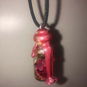 Puede incluir: Un pequeño colgante de botella de vidrio con un sello de cera roja y brillante. La botella contiene flores rojas secas y escamas doradas. El colgante está unido a un cordón negro.