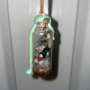 May include: A small glass bottle filled with various crystals, glitter, and small stars. The bottle is sealed with a green wax-like substance and hangs from a brown cord.