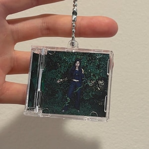 Puede incluir: Un llavero rectangular transparente con una cadena de bolas plateada. El llavero contiene una pequeña imagen de una persona vestida de oscuro frente a un fondo verde y frondoso.