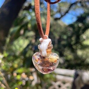 May include: A heart-shaped glass bottle filled with dried flowers and herbs, hanging from a brown leather cord. The bottle has a white wax seal on the top.