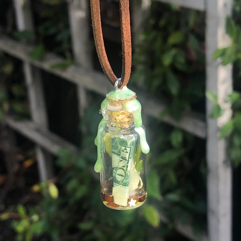 Puede incluir: Un collar con una botella de vidrio verde, un tap&oacute;n de corcho y un billete de d&oacute;lar en su interior. La botella est&aacute; llena de un l&iacute;quido transparente y tiene una sustancia verde cerosa que gotea por los lados.