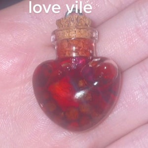 May include: A heart-shaped glass bottle filled with a red liquid and small pieces of gold glitter. The bottle has a cork stopper and a silver chain.
