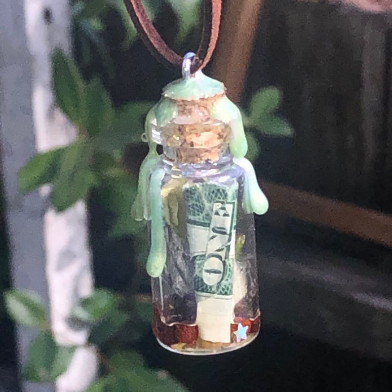 Puede incluir: Un collar con una peque&ntilde;a botella de vidrio sellada con cera verde y un billete de d&oacute;lar enrollado en su interior. La botella est&aacute; llena de un l&iacute;quido transparente y tiene un tap&oacute;n de corcho.