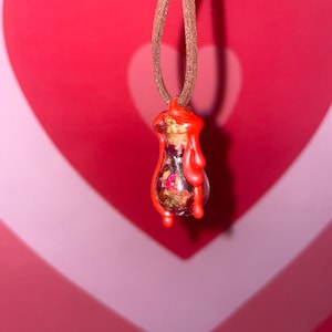 May include: A red glass bottle filled with dried flowers and sealed with red wax hangs from a brown cord. The bottle is shaped like a teardrop and has a gold lid.