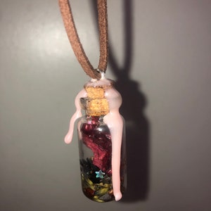 Puede incluir: Una pequeña botella de vidrio con un tapón de corcho y un sello de cera rosa. La botella está llena de hierbas secas, flores y purpurina. Está colgada de un cordón marrón.