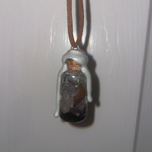May include: A silver-painted glass bottle filled with various crystals and stones, hanging from a brown leather cord. The bottle has a cork stopper and is sealed with a silver-colored wax.