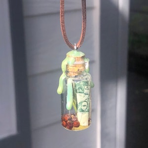 Puede incluir: Un collar con un colgante de botella de vidrio peque&ntilde;a con un tap&oacute;n de corcho. La botella est&aacute; llena de cera verde, hojas secas y peque&ntilde;as semillas marrones. El collar est&aacute; hecho con un cord&oacute;n de cuero marr&oacute;n.