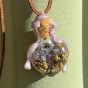 May include: A clear glass pendant in the shape of a heart, filled with dried herbs and crystals, and sealed with a cork. The pendant is encased in a lavender-colored resin and hangs from a brown leather cord.