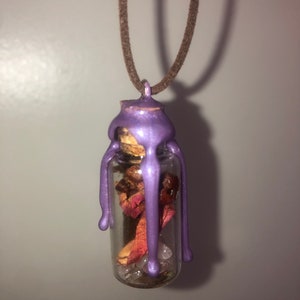 Peut inclure: Un collier avec un flacon en verre scell&eacute; &agrave; la cire violette et un bouchon en li&egrave;ge. Le flacon contient des p&eacute;tales de rose s&eacute;ch&eacute;es et des cristaux transparents.