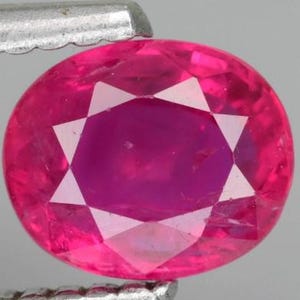 Könnte beinhalten: Ein leuchtender, oval geschliffener Edelstein in einem tiefen Pink-Rot mit einer facettierten Oberfläche, die in der Mitte ein sternförmiges Muster bildet. Der Stein wird sicher zwischen den gezackten Metallspitzen einer Pinzette gehalten.