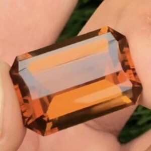 May include: A rectangular, faceted gemstone with a warm, amber-orange hue. The gem is held between fingers, showcasing its clarity and multiple facets. The cut of the gem is an emerald cut, with a rectangular shape and beveled corners.