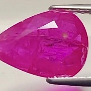 Puede incluir: Una gema facetada en forma de lágrima, de un vibrante color magenta. La gema está sujeta por unas pinzas plateadas, mostrando sus superficies pulidas y su claridad interna. El corte de la gema revela múltiples facetas.