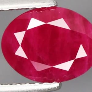 May include: A faceted, oval-shaped ruby gemstone. The ruby is a deep, saturated red color with white triangular facets. The gemstone is held by a silver metal tool.