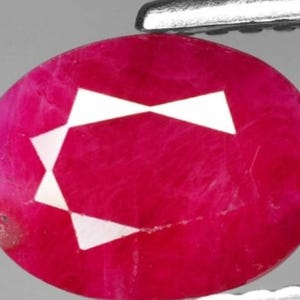 May include: Oval-cut ruby gemstone with a vibrant red hue. The gemstone features a unique white geometric pattern in the center. Held by a silver tool, the ruby is a striking example of natural beauty.