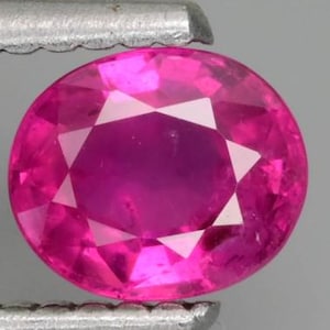 Pode incluir: Uma gema de corte oval vibrante com uma cor rosa choque profunda e saturada. A pedra apresenta uma superfície facetada que reflete a luz, criando uma aparência brilhante e cintilante. É mantida por uma ferramenta metálica de borda serrilhada.