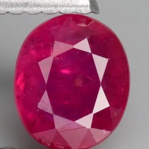 Pode incluir: Uma gema de rubi de corte oval, com uma cor vermelha profunda e saturada e uma superfície multifacetada. A pedra é mantida por uma ferramenta metálica de bordas serrilhadas na parte superior e inferior, exibindo sua clareza e facetas geométricas.