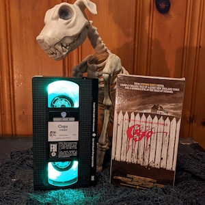 Puede incluir: Una cinta VHS de la película de terror Cujo, con una imagen en blanco y negro de un perro con sangre roja goteando de su boca. La cinta está en una caja negra con una luz verde brillando a través de la ventana. La caja está sentada sobre una superficie negra con una figura de perro esqueleto en el fondo.