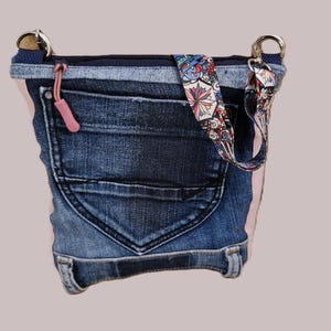 Könnte beinhalten: Eine Jeans-Umhängetasche mit einem rosa Akzentstreifen und einem gemusterten Riemen. Die Tasche hat eine Vordertasche und einen rosa Reißverschlussanhänger. Die Tasche besteht aus recyceltem Denim.
