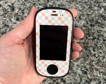 May include: A small, handheld device with a black screen and a colorful checkered pattern on the front. The device has a black border and a circular button at the bottom. The checkered pattern includes pastel shades of pink, orange, and blue.