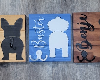 May include: Three wooden dog leash hooks with black metal hooks. The hooks are shaped like dog butts and have the names Lassie, Buster, and Benny written on them. The hooks are painted in different colors: brown, blue, and light brown.