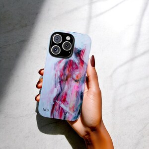 以下が含まれることがあります： カラフルな抽象的な人物の胴体の絵が描かれた白い携帯電話ケース。絵はピンク、赤、青の色合いで描かれています。ケースにはカメラレンズの切り抜きと、下部に署名があります。